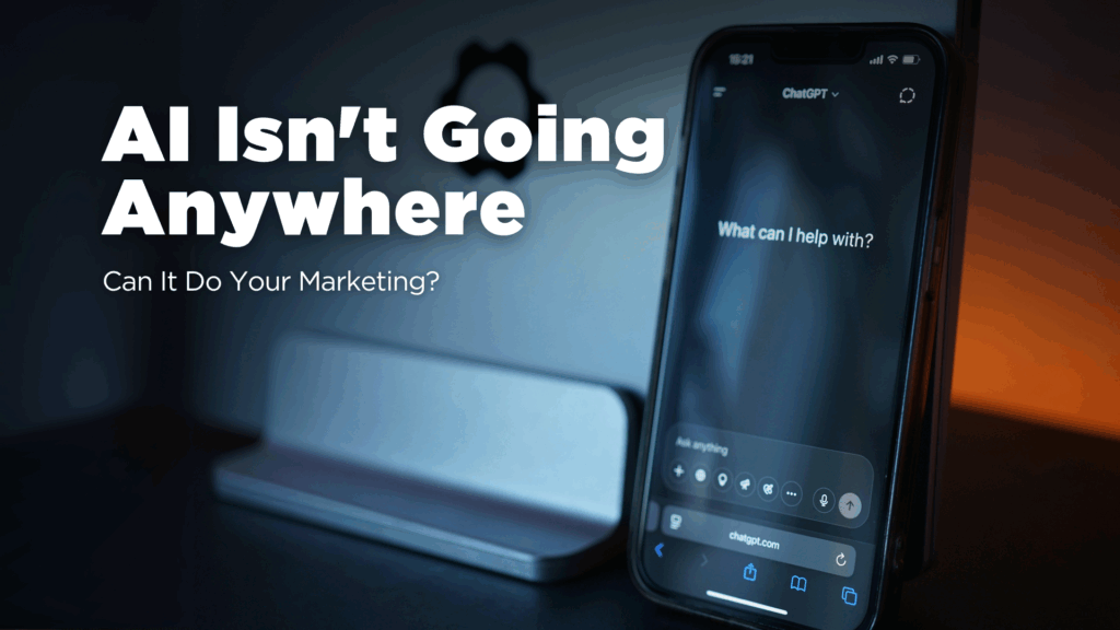 A dark, moody image featuring a smartphone displaying the ChatGPT interface ("What can I help with?") propped against a silver laptop stand. White text overlaid on the image reads: "AI Isn't Going Anywhere" and "Can It Do Your Marketing?"